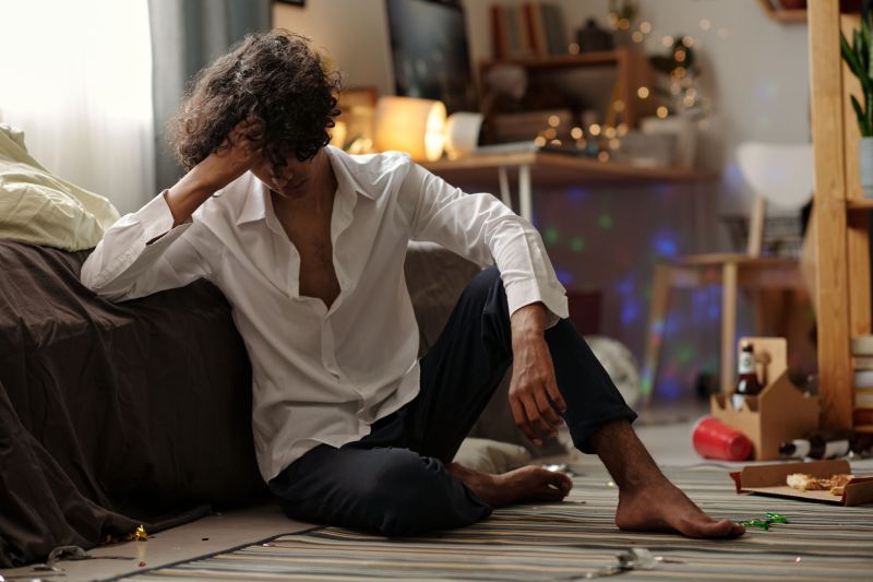 Young man with dark curly hair touching his head while suffering from symptoms of hangover in the morning after home party